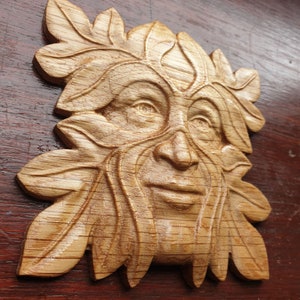 K&ouml;nnte beinhalten: Eine Holzschnitzerei eines Green Man-Gesichts, mit Bl&auml;ttern und &Auml;sten, die sein Haar und seinen Bart bilden. Die Schnitzerei ist detailliert und realistisch, mit einer warmen braunen Farbe.