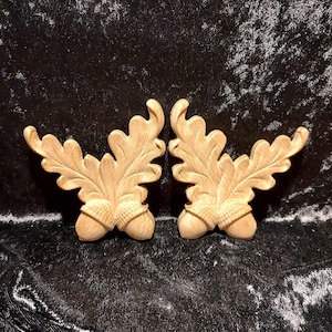 May include: Two wooden acorn and oak leaf ornaments. The ornaments are carved in a detailed, realistic style. They are a light brown colour and have a smooth, polished finish.