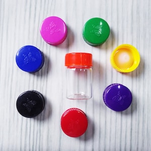 May include: A clear glass vial with a bright orange screw-top lid, surrounded by seven additional plastic bottle caps in various colors including pink, green, yellow, purple, red, blue, and black, each featuring a small embossed butterfly design.