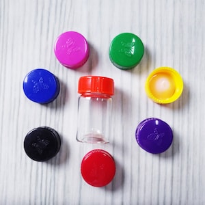 May include: A clear glass vial with a red screw-top lid, surrounded by seven individual plastic bottle caps in various colors including pink, green, yellow, purple, red, blue, and black, each featuring a small embossed butterfly design.