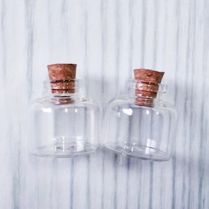 May include: Two clear glass bottles with cork stoppers. The bottles are empty and have a round shape.
