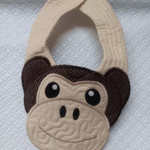 May include: A beige and brown monkey-faced baby bib. The bib features a monkey face with large black eyes, a smiling mouth, and brown polka-dot ears. The bib has a snap closure at the neck and is made of soft fabric.