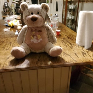 May include: A plush brown and white teddy bear with a pink shirt that says "Melina" on it. The bear is sitting on a wooden table.