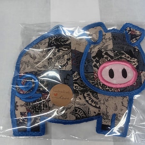 May include: A handmade pig-shaped applique, with a patchwork design in blue, black, and beige. The pig has a pink nose and blue stitched outline. A small, round sticker with the words "handmade with love" is visible.