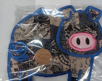 Op de afbeelding: Een handgemaakte varkensvormige applicatie, met een patchwork-ontwerp in blauw, zwart en beige. Het varken heeft een roze neus en een blauwe gestikte omtrek. Een kleine, ronde sticker met de tekst "handmade with love" is zichtbaar.