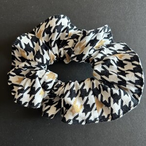 Peut inclure: Un chouchou en tissu avec un motif pied-de-poule noir, blanc et doré. L'accessoire pour cheveux est froncé et élastique, créant un effet volanté. Parfait pour attacher les cheveux en chignon ou en queue de cheval.