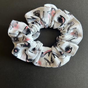 May include: White scrunchie featuring a repeating pattern of high heels in black, grey, and pink. The fabric is gathered and elasticated, creating a ruffled look. A fashionable hair accessory.