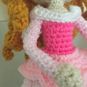 Puede incluir: Una mu&ntilde;eca de crochet con el pelo largo, rizado y dorado, que lleva un vestido rosa con un cuello blanco y un brazo blanco de crochet.