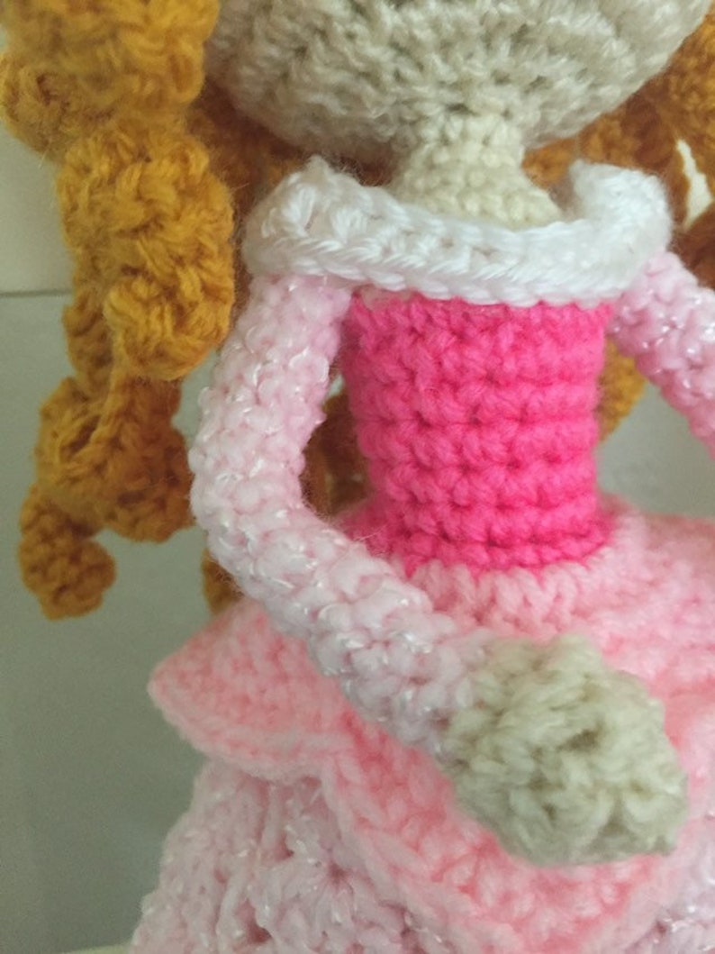 Puede incluir: Una mu&ntilde;eca de crochet con el pelo largo, rizado y dorado, que lleva un vestido rosa con un cuello blanco y un brazo blanco de crochet.