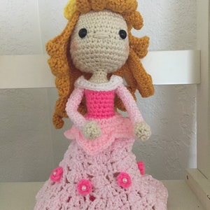 Puede incluir: Una mu&ntilde;eca de crochet con un vestido rosa y una corona amarilla. La mu&ntilde;eca tiene el pelo largo y casta&ntilde;o y los ojos negros.