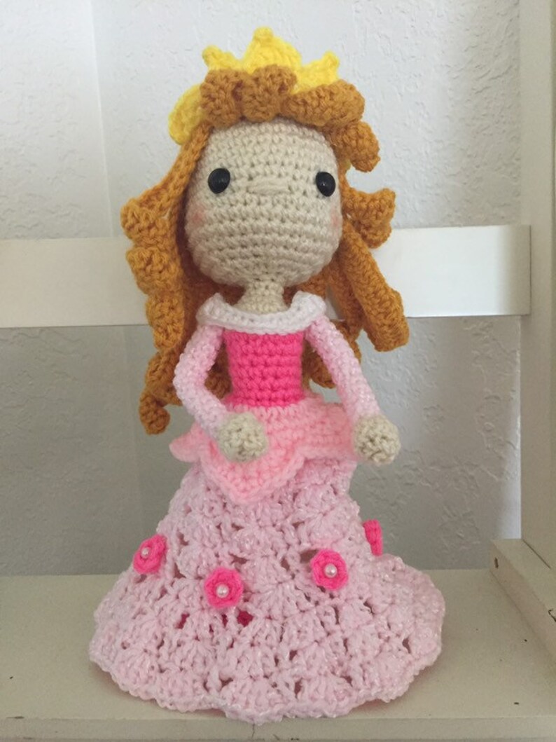 Puede incluir: Una mu&ntilde;eca de crochet con un vestido rosa y una corona amarilla. La mu&ntilde;eca tiene el pelo largo y casta&ntilde;o y los ojos negros.