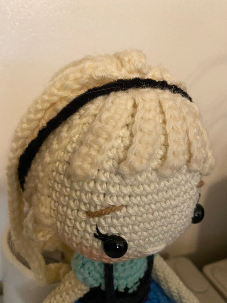 K&ouml;nnte beinhalten: Eine geh&auml;kelte Puppe mit blonden Haaren, schwarzen Augen und einem schwarzen Stirnband. Die Puppe tr&auml;gt ein blaues und gr&uuml;nes Kleid.