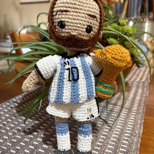 May include: A crocheted doll featuring a brown-haired figure with a beard, wearing a white and light blue striped soccer jersey with the number 10 on the chest and shorts. The doll holds a golden trophy with green accents in one hand.