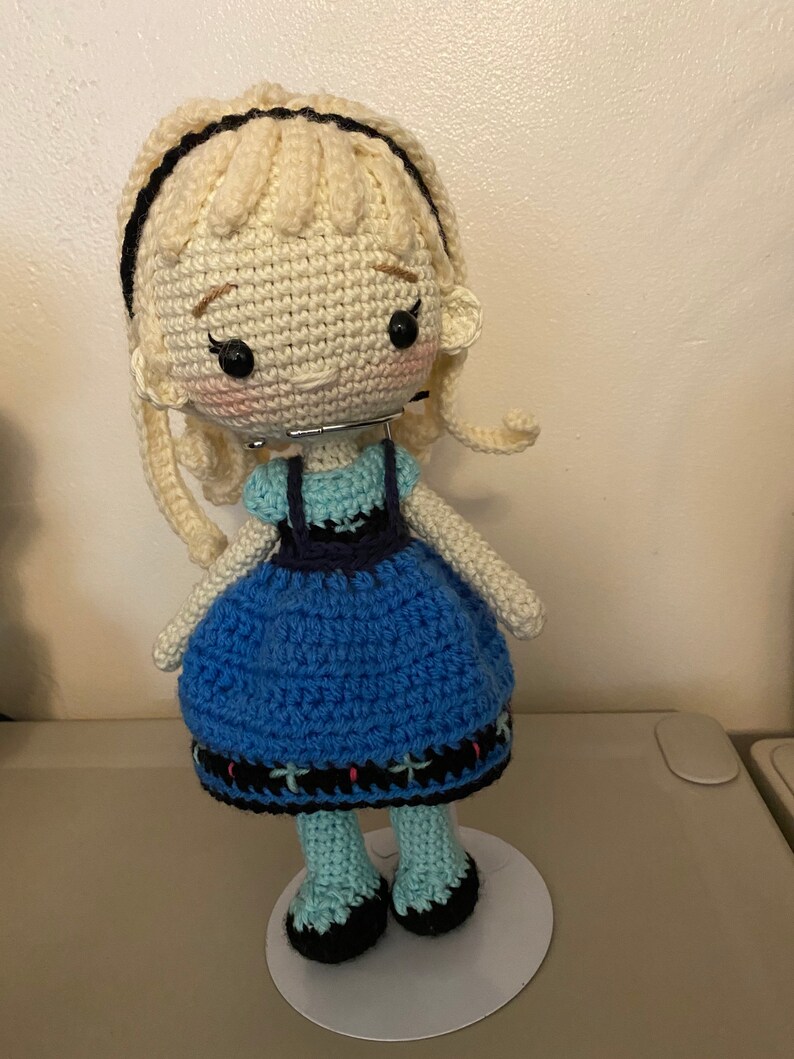 K&ouml;nnte beinhalten: Eine geh&auml;kelte Puppe mit blonden Haaren, einem blauen Kleid und schwarzen Schuhen. Die Puppe steht auf einem wei&szlig;en Sockel.