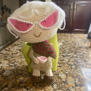 May include: A crocheted doll with white hair, pink sunglasses, and a brown scarf holding a pink cup. The doll is sitting on a green planter.