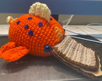 May include: A crocheted orange and brown plush toy shaped like a fish with a blue eye. The fish is holding a brown crocheted sandwich in its mouth.