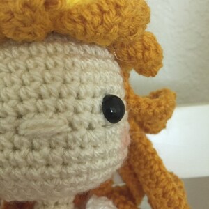 Puede incluir: Una mu&ntilde;eca de crochet con una corona amarilla y un vestido blanco. La mu&ntilde;eca tiene un ojo negro y un rubor rosado en las mejillas.