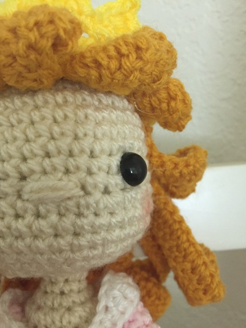 Puede incluir: Una mu&ntilde;eca de crochet con una corona amarilla y un vestido blanco. La mu&ntilde;eca tiene un ojo negro y un rubor rosado en las mejillas.
