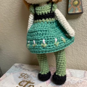 K&ouml;nnte beinhalten: H&auml;kelpuppe mit braunen Haaren, einem gr&uuml;nen Kleid mit schwarzem Besatz und schwarzen Schuhen. Die Puppe steht auf einer Oberfl&auml;che mit dem Aufdruck "to travel is to".