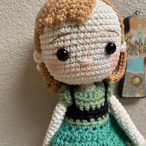 K&ouml;nnte beinhalten: H&auml;kelpuppe mit braunen Haaren, wei&szlig;er Haut und schwarzen Augen. Die Puppe tr&auml;gt ein gr&uuml;nes Kleid mit einem schwarzen Oberteil.