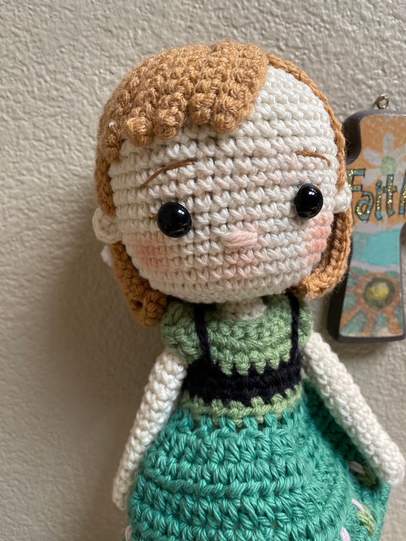 K&ouml;nnte beinhalten: H&auml;kelpuppe mit braunen Haaren, wei&szlig;er Haut und schwarzen Augen. Die Puppe tr&auml;gt ein gr&uuml;nes Kleid mit einem schwarzen Oberteil.