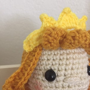 Puede incluir: Una mu&ntilde;eca de crochet con una corona amarilla y cabello marr&oacute;n. La mu&ntilde;eca tiene ojos negros y una cara blanca.
