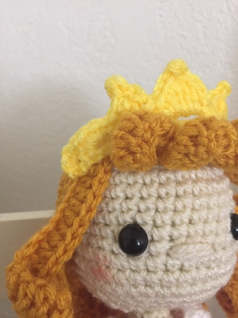 Puede incluir: Una mu&ntilde;eca de crochet con una corona amarilla y cabello marr&oacute;n. La mu&ntilde;eca tiene ojos negros y una cara blanca.