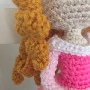 Puede incluir: Una mu&ntilde;eca de crochet con el pelo largo, rubio dorado, que lleva un vestido rosa con ribete blanco. La cara de la mu&ntilde;eca es blanca con mejillas rosadas.
