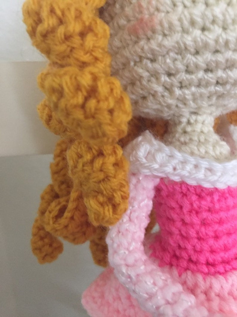 Puede incluir: Una mu&ntilde;eca de crochet con el pelo largo, rubio dorado, que lleva un vestido rosa con ribete blanco. La cara de la mu&ntilde;eca es blanca con mejillas rosadas.