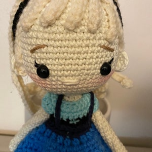 K&ouml;nnte beinhalten: Eine geh&auml;kelte Puppe mit blonden Haaren, einem blauen Kleid und schwarzen Augen. Die Puppe tr&auml;gt ein blaues und wei&szlig;es H&auml;kelkleid mit einem schwarzen H&auml;kelmieder.