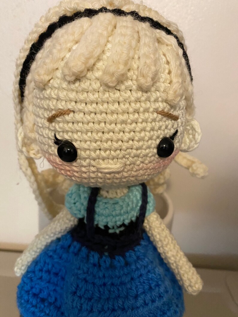 K&ouml;nnte beinhalten: Eine geh&auml;kelte Puppe mit blonden Haaren, einem blauen Kleid und schwarzen Augen. Die Puppe tr&auml;gt ein blaues und wei&szlig;es H&auml;kelkleid mit einem schwarzen H&auml;kelmieder.