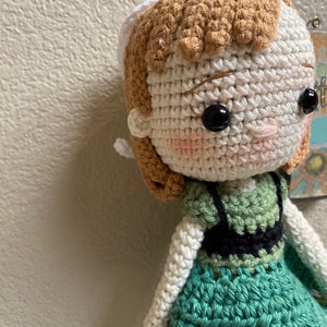 K&ouml;nnte beinhalten: H&auml;kelpuppe mit braunen Haaren, einer wei&szlig;en H&auml;kelm&uuml;tze und einem gr&uuml;nen Kleid. Die Puppe hat schwarze Augen und rosafarbene Wangen.