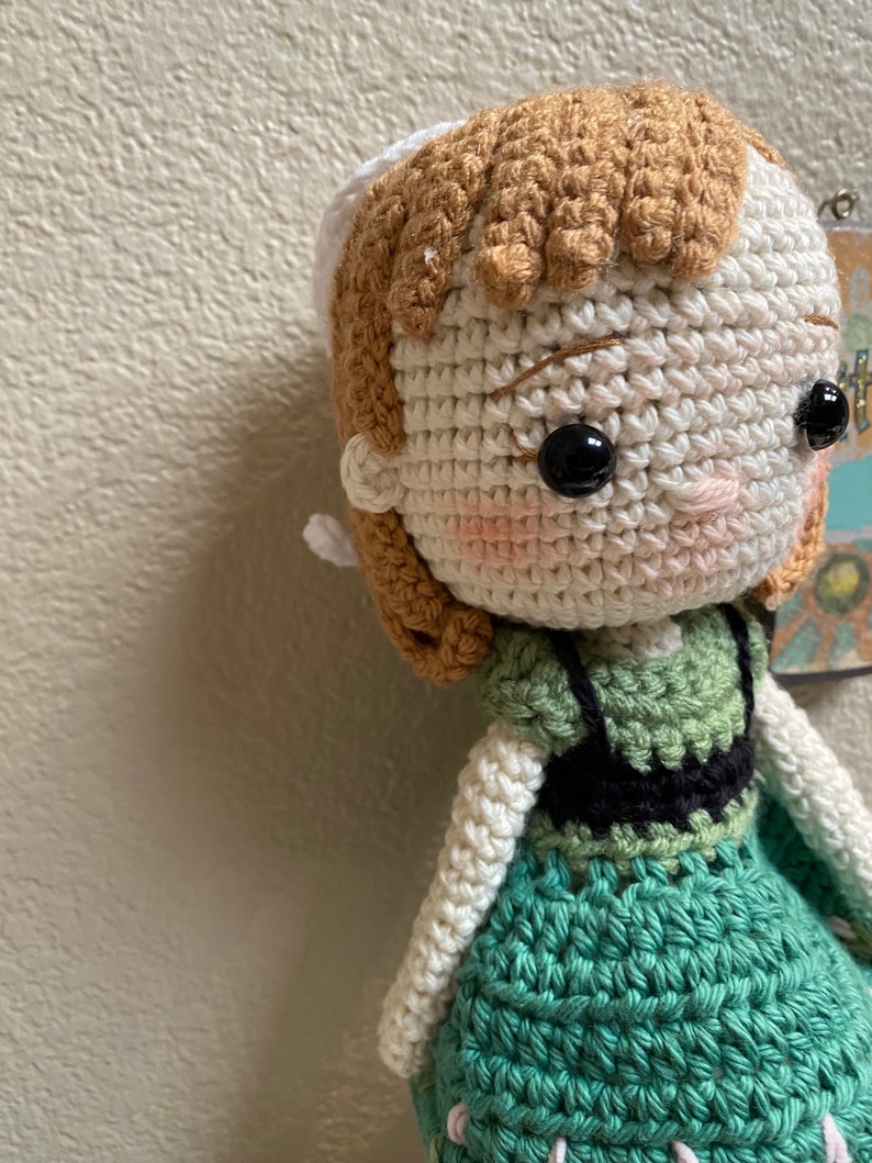 K&ouml;nnte beinhalten: H&auml;kelpuppe mit braunen Haaren, einer wei&szlig;en H&auml;kelm&uuml;tze und einem gr&uuml;nen Kleid. Die Puppe hat schwarze Augen und rosafarbene Wangen.