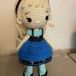 K&ouml;nnte beinhalten: Eine geh&auml;kelte Puppe mit blonden Haaren, einem blauen Kleid und schwarzen Schuhen. Die Puppe tr&auml;gt ein Stirnband und hat einen s&uuml;&szlig;en Gesichtsausdruck.