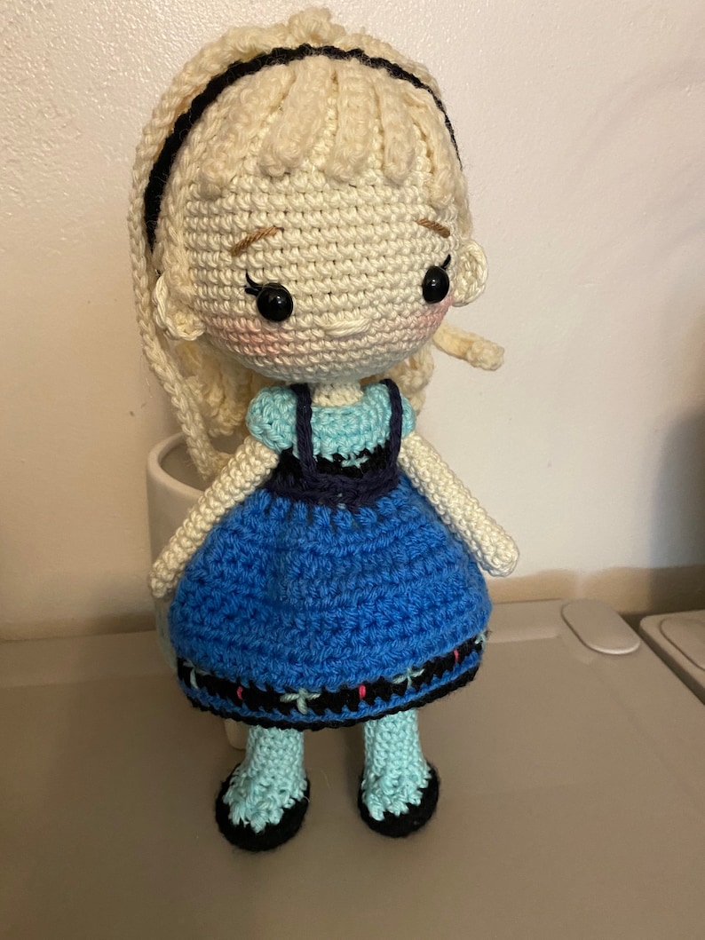 K&ouml;nnte beinhalten: Eine geh&auml;kelte Puppe mit blonden Haaren, einem blauen Kleid und schwarzen Schuhen. Die Puppe tr&auml;gt ein Stirnband und hat einen s&uuml;&szlig;en Gesichtsausdruck.