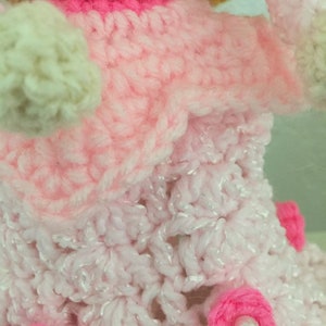 Puede incluir: Una mu&ntilde;eca de crochet con un vestido rosa, una falda blanca y flores rosas. La mu&ntilde;eca est&aacute; hecha con una variedad de hilos rosas y blancos.