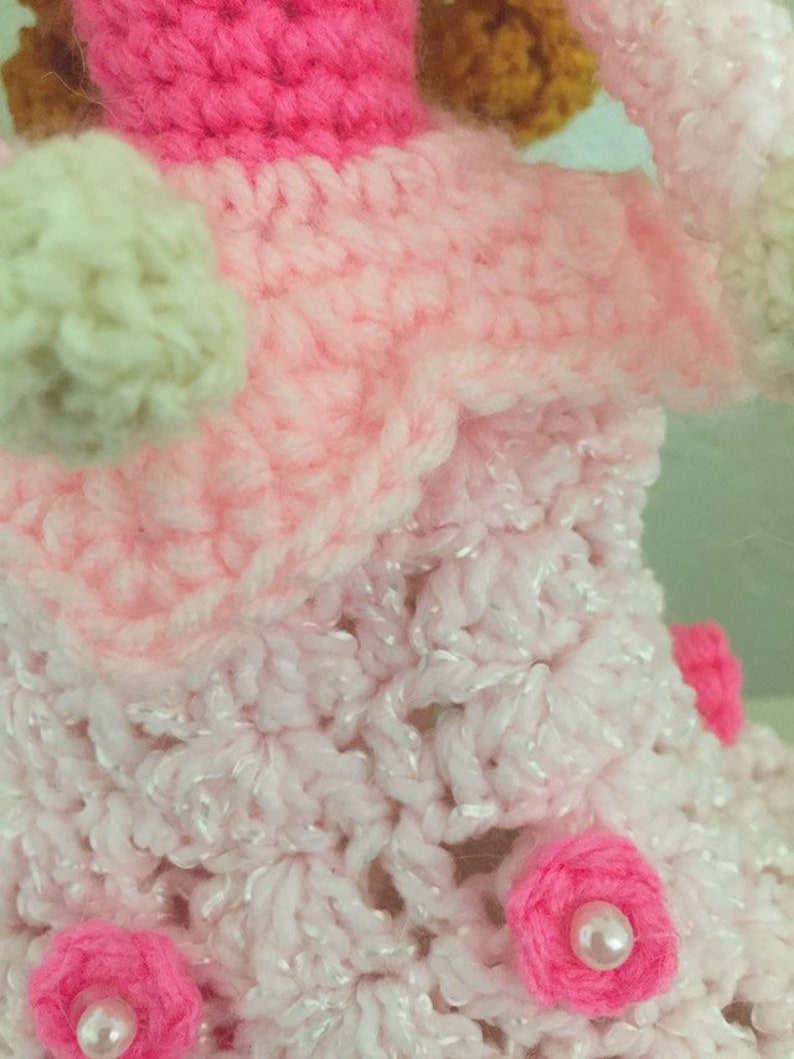 Puede incluir: Una mu&ntilde;eca de crochet con un vestido rosa, una falda blanca y flores rosas. La mu&ntilde;eca est&aacute; hecha con una variedad de hilos rosas y blancos.