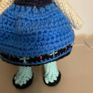 K&ouml;nnte beinhalten: Eine geh&auml;kelte Puppe, die ein blaues Kleid mit schwarzem und wei&szlig;em Besatz tr&auml;gt. Die Puppe hat wei&szlig;es Garnhaar und tr&auml;gt blaue und schwarze Schuhe.