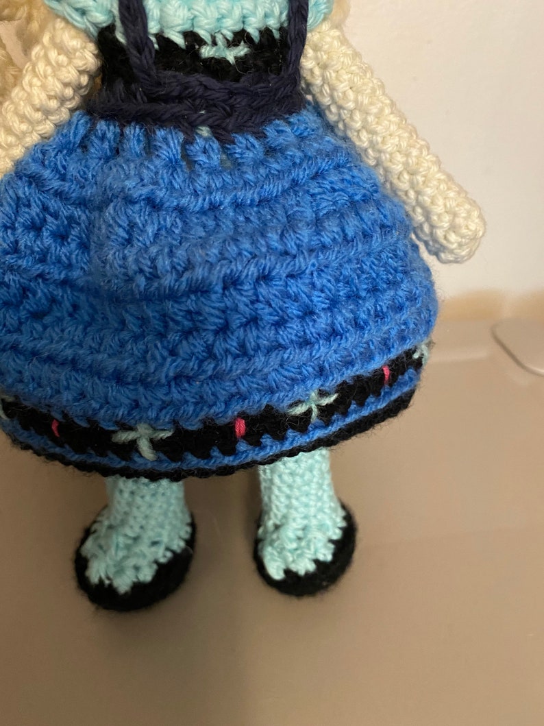 K&ouml;nnte beinhalten: Eine geh&auml;kelte Puppe, die ein blaues Kleid mit schwarzem und wei&szlig;em Besatz tr&auml;gt. Die Puppe hat wei&szlig;es Garnhaar und tr&auml;gt blaue und schwarze Schuhe.