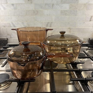 May include: A set of three amber-colored glass cookware pieces, including a large stockpot, a medium saucepan with a lid, and a wide casserole dish with a lid. Each piece features integrated handles and matching glass lids with dark brown knobs.