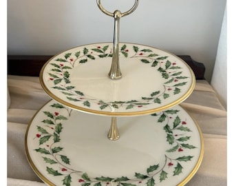 May include: A two-tier serving stand featuring white ceramic plates with a green holly leaf and red berry pattern along the rims. The plates are accented with gold trim and connected by a central polished gold-tone metal handle with a loop top.