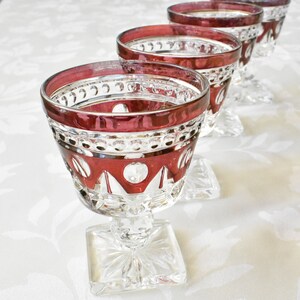 May include: Set of four clear glass stemmed drinking glasses with a red rim and intricate cut glass design. The glasses have a square base.