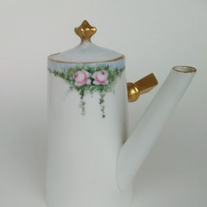 Puede incluir: Tetera de porcelana blanca con tapa y asa doradas. La tetera tiene un delicado diseño floral con rosas rosas y hojas verdes.