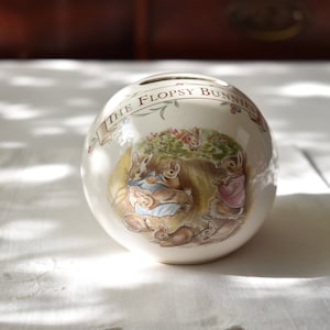 May include: A white ceramic piggy bank shaped like an egg with a colorful illustration of Peter Rabbit and his siblings. The bank has the text "The Flopsy Bunnies" on the front.