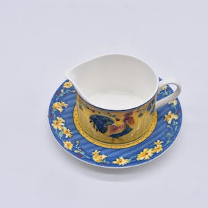May include: A white ceramic gravy boat with a handle, sitting on a matching saucer. The saucer and the boat's base are yellow with a blue rooster design. The saucer has a blue background with yellow floral accents.