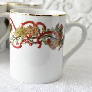 May include: White ceramic mug with a gold rim and a festive holiday design featuring red ribbons, green holly leaves, and red berries.