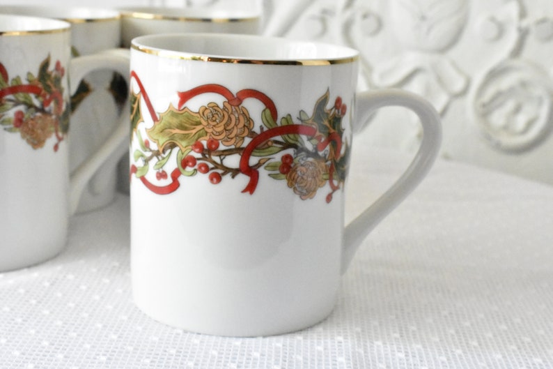 May include: White ceramic mug with a gold rim and a festive holiday design featuring red ribbons, green holly leaves, and red berries.