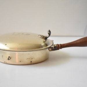 May include: A silver-colored, lidded pan with a wooden handle. The pan has a decorative handle on the lid and a detailed rim. The handle is a dark brown color. The pan is on a white surface.