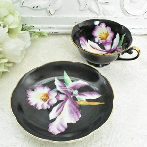 May include: A black teacup and saucer set with a floral design. The cup and saucer are black with a gold trim. The floral design features purple and white orchids. The set is displayed on a white lace tablecloth.
