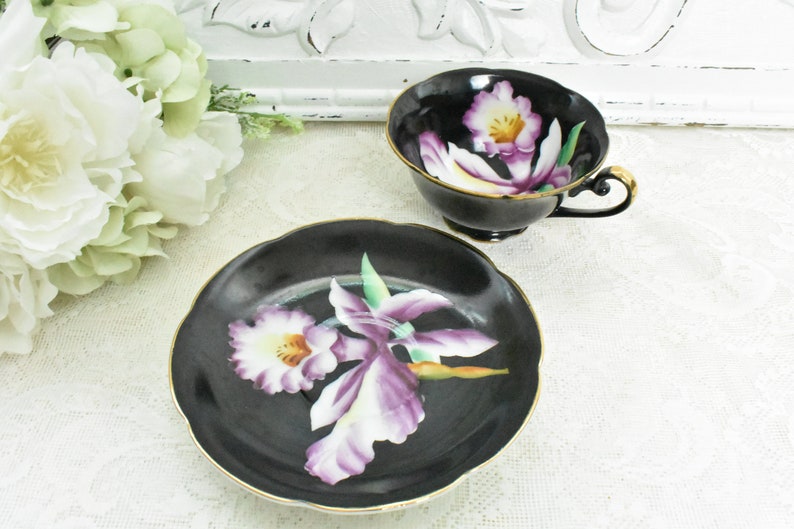 May include: A black teacup and saucer set with a floral design. The cup and saucer are black with a gold trim. The floral design features purple and white orchids. The set is displayed on a white lace tablecloth.